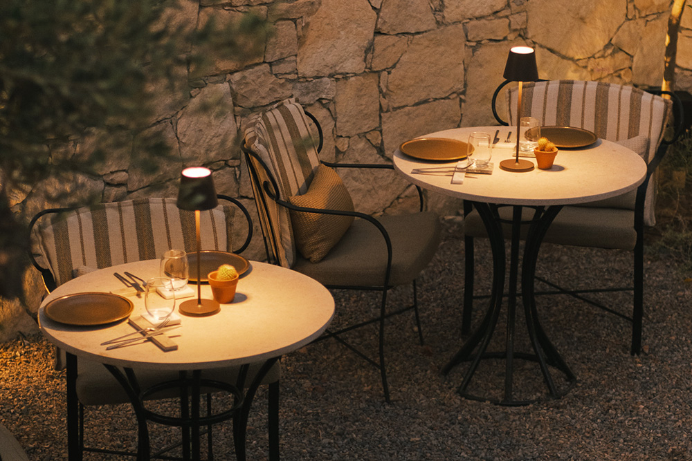 The Olive Grove at Elia at MOMI, small stone tables and wrought-iron chairs set against a dry-stone wall lit by table lamps at dusk
