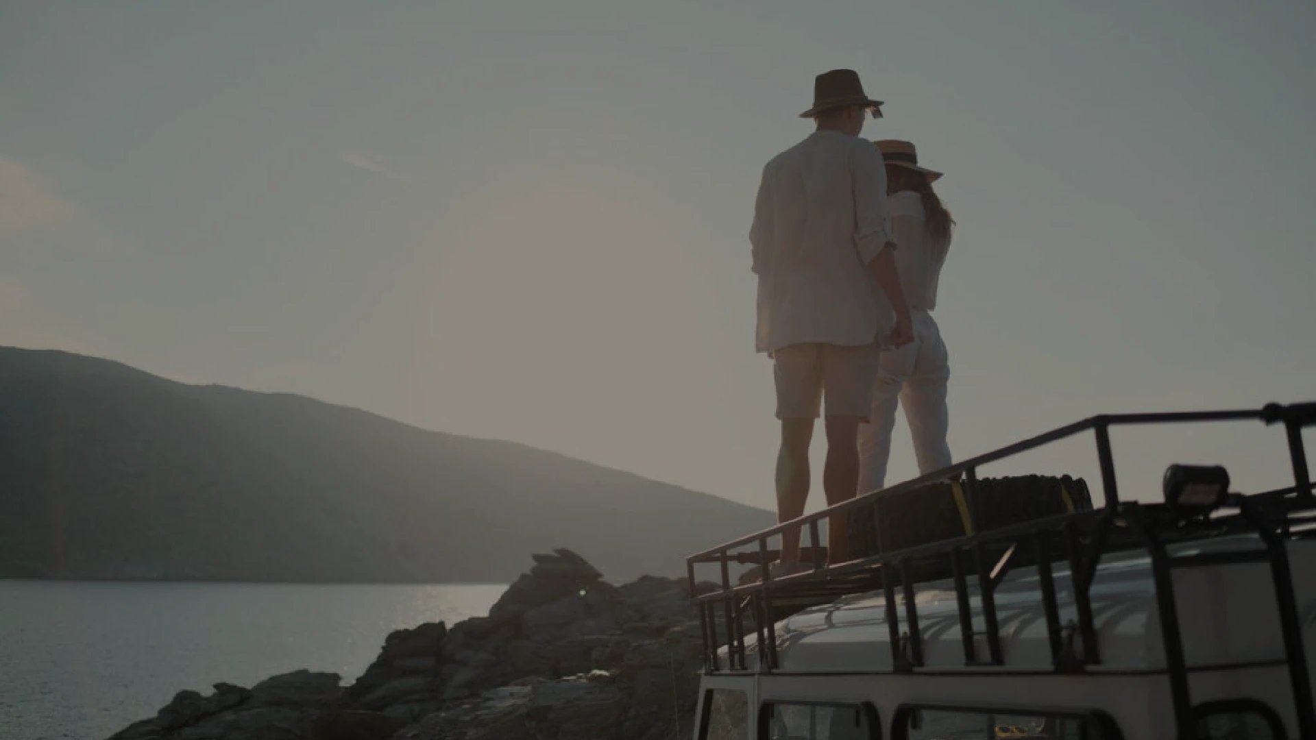 Couple standing on a jeep roof rack at sunset by the sea