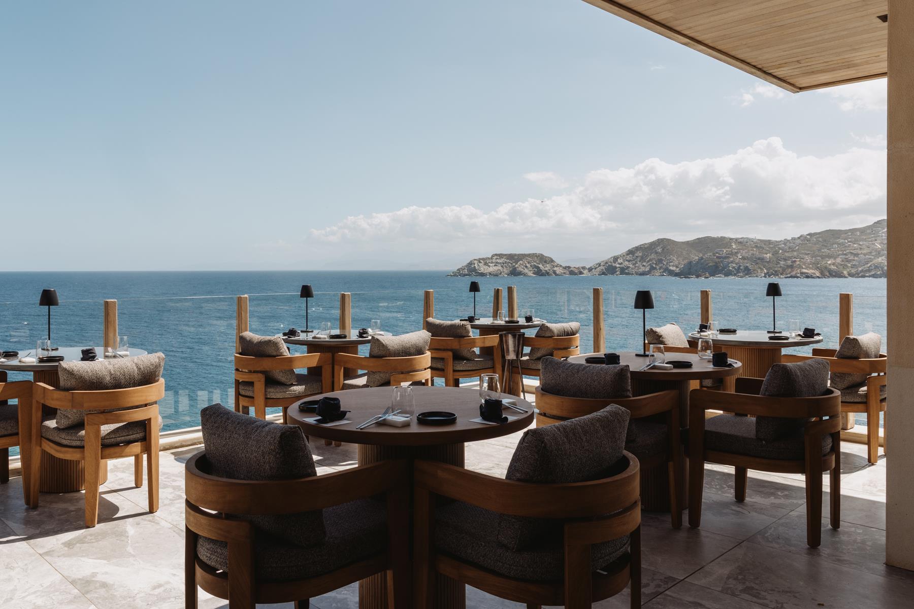 Oceano restaurant at Seaside, round timber tables with dark lamps on a marble terrace facing the sea and distant Cretan headlands