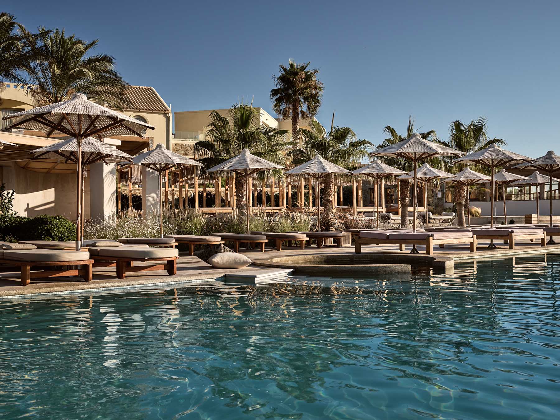 The poolside lounge at Seaside, woven umbrellas, sun loungers and palm trees reflected in the calm pool water