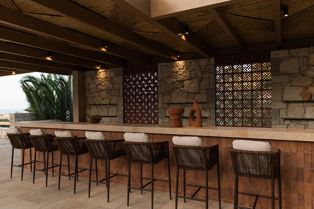 The pool bar at MOMI, a long timber counter beneath a reed-lined pergola with woven barstools