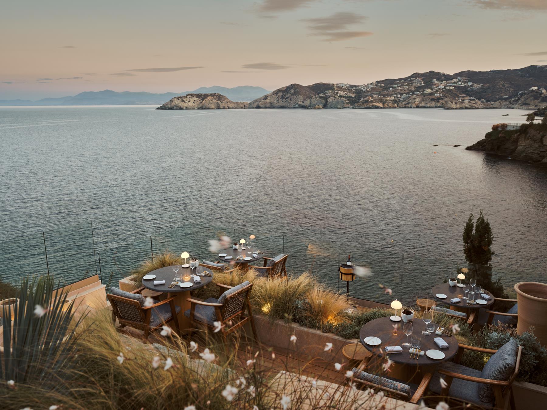 Caléa at Seaside, terrace tables set for dinner above the bay at twilight, glass railings and the Cretan Sea beyond