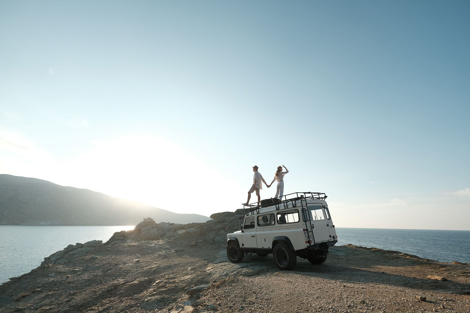 A vintage off-road vehicle parked on a rocky headland at golden hour, two people looking out over the coast